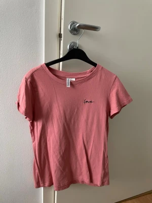 Rosa t-shirt med broderad text - Säljer en enkel och snygg rosa t-shirt från H&M med liten broderad text 'love' på bröstet. T-shirten har rund halsringning och korta ärmar. Perfekt till jeans eller kjol för en avslappnad look.