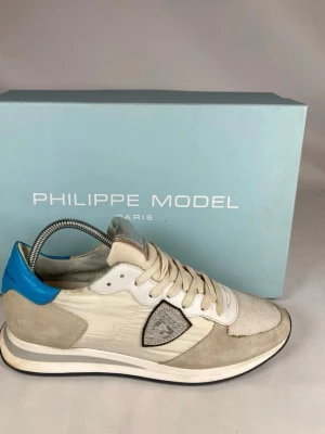 Philippe Model  - Snygga sneakers från Philippe Model i beige och vitt med detaljer i silver och blått. Skorna har snörning, mocka- och skinnpaneler samt en klassisk sportig siluett. Perfekta för dig som vill ha en stilren men unik sneaker.