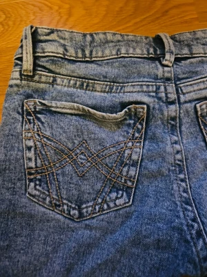 Blå bootcut jeans från gina tricot - Säljer ett par blå jeans med bootcut  och snygga broderade detaljer på bakfickorna. Jeansen har klassisk femficksdesign och bälteshällor. Materialet är denim i 100% bomull och färgen är tvättad blå. Perfekta för en avslappnad och trendig look.