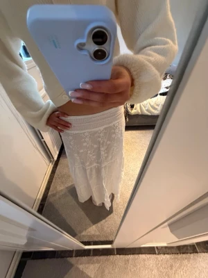 Vit långkjol med spetsdetaljer - Säljer en romantisk vit långkjol med fina spetsdetaljer och broderade blommor. Kjolen har bred resår i midjan och faller snyggt längs benen. Perfekt för dig som gillar en bohemisk och somrig stil.