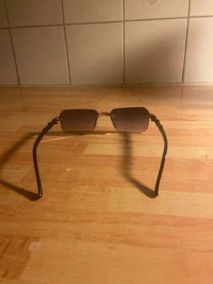 Bruna rektangulära solglasögon - Snygga solglasögon med rektangulära, bruna glas och gulddetaljer vid bågarna. Glasen är utan synlig ram och skalmarna är svarta. Perfekta för dig som vill ha en stilren och modern look.