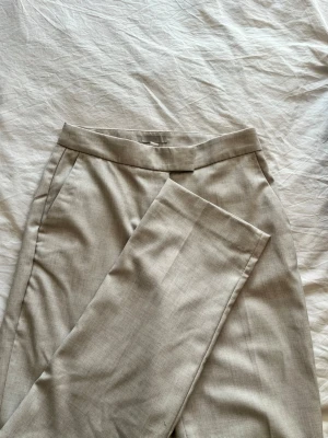 Beige kostymbyxa med raka ben - Stilrena beige kostymbyxor med hög midja och raka ben. Byxorna har diskreta sidofickor och bred linning med dold knäppning.