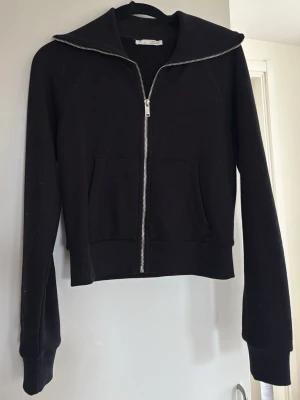 Svart zip hoodie med fickor - Snygg svart hoodie med dragkedja framtill och två stora fickor. Hoodien har en bred krage och ribbade muddar vid ärmslut och nederkant. Perfekt för en avslappnad och cool stil. Materialet känns mjukt och bekvämt.