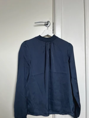 Mörkblå satinblus  - Snygg mörkblå blus i satinliknande material med långärmad modell. Blusen har resårdetaljer vid halsringningen. Den är endast använd en gång så är i fint skick!