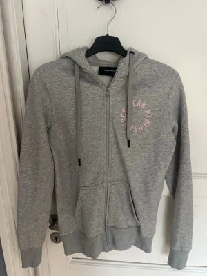 Grå zip-hoodie från Peak Performance - Snygg grå hoodie från Peak Performance med dragkedja, huva med dragsko och känguruficka. Rosa logga tryckt på bröstet. Använd få gånger.