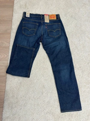 Levi's blå raka jeans - Säljer ett par klassiska blå jeans från Levi's med rak passform och fem fickor. Jeansen har snygga slitningar framtill och kontrastsömmar. Tillverkade i slitstark denim med Levi's ikoniska läderpatch bak i midjan.