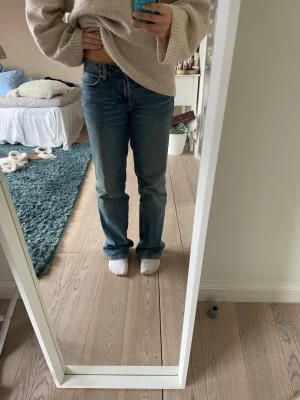 Blå raka jeans med låg midja - Säljer ett par klassiska blå jeans med rak passform och låg midja. Jeansen har fem fickor, bälteshällor och stängs med knapp och dragkedja. Materialet är denim i bomull och färgen är mellanblå med lätt slitning på låren. Lite slitna längst nere