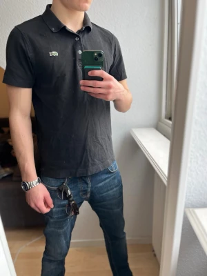 Svart pikétröja från Lacoste - Klassisk svart pikétröja från Lacoste med den ikoniska gröna krokodilloggan på bröstet. Tröjan har korta ärmar, krage och knappar framtill. Tillverkad i mjuk bomull som känns skön mot huden. Perfekt till jeans eller shorts för en clean look.