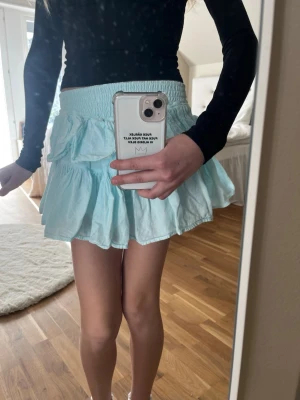 Ljusblå volangkjol med resårmidja - Supersöt ljusblå kortkjol med volanger och bred resår i midjan. Kjolen har en luftig och flowy känsla, perfekt för varmare dagar. Passar till både enkla toppar och stickade tröjor för en trendig look.