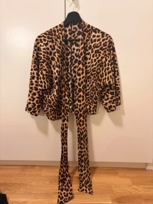 Kimono med leopardmönster - Säljer en snygg kimono med leopardmönster i brunt, svart och beige. Modellen har vida ärmar och ett matchande knytband i midjan. Perfekt för att addera lite edge till din outfit.
