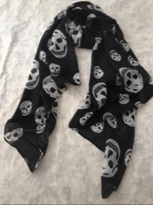 Svart scarf med dödskallemönster - Cool svart halsduk med vita dödskallar i olika storlekar. Tunt och lätt material som ger en edgy vibe till din outfit. Perfekt att bära runt halsen eller som sjal. Passar dig som gillar alternativ stil och vill sticka ut.