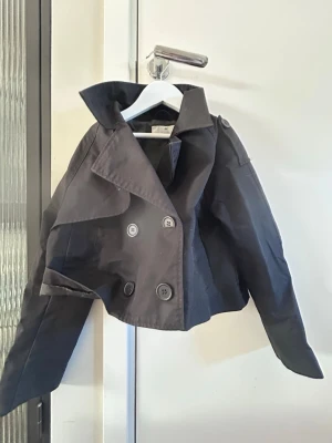 Svart kort trenchcoat från H&M - Stilren svart trenchcoat från H&M i kort modell med dubbelknäppning och bred krage. Jackan har axelklaffar och är tillverkad i ett lätt tyg som passar perfekt till lager på lager. Snygg detalj med stora knappar framtill.