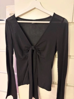 Svart långärmad topp med knyt - Trendig svart långärmad topp med djup v-ringning och knyt framtill. Tunn och lätt i materialet, perfekt för lager på lager eller som statement-plagg. Snygg passform som framhäver midjan och ger en stilren look.