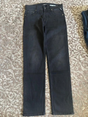 Svarta raka jeans från Replay - Säljer ett par svarta jeans från Replay med rak passform och klassisk femficksdesign. Jeansen har diskreta sömmar och metallknappar. Materialet är denim med en mjuk känsla och lätt stretch. Perfekta för dig som gillar en clean och stilren look.
