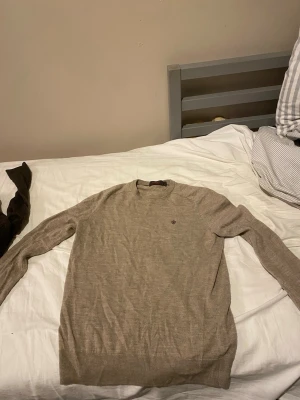 Beige stickad Morris tröja med rund hals - Snygg beige stickad tröja med rund halsringning och diskret broderad logga på bröstet. Tröjan har långa ärmar och ribbade muddar vid ärmslut och nederkant. Perfekt för lager på lager och enkel att matcha med olika outfits. Äkta Morris! Säljs för den är för liten