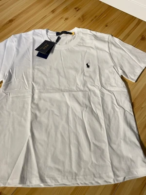 Vit t-shirt från Polo Ralph Lauren - Klassisk vit t-shirt från Polo Ralph Lauren med rund hals och den ikoniska lilla broderade loggan på bröstet. Tillverkad i mjuk bomull för en skön känsla och perfekt passform. Enkel och stilren design som funkar till allt.