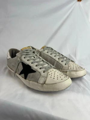 Golden Goose Superstar Skor - Snygga vita sneakers från Golden Goose med grå meshdetaljer och en ikonisk svart stjärna på sidan. Skorna har perforerad tå, grå skosnören och gulddetalj på plösen. Perfekta för en trendig och avslappnad streetstyle-look.
