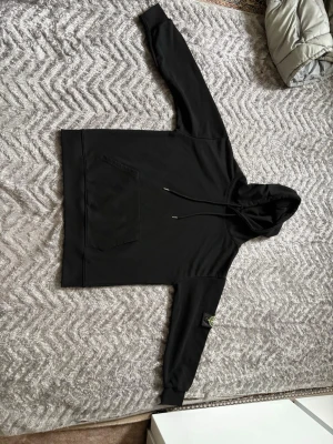 Svart hoodie från Stone Island - Säljer en svart hoodie från Stone Island med klassisk huva och dragsnören. Hoodien har en stor magficka framtill och den ikoniska Stone Island-loggan på vänster ärm. Tillverkad i mjukt bomullsmaterial och har ribbade muddar vid ärmslut och nederkant.