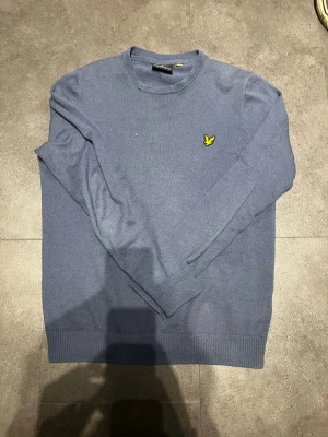 Blå stickad tröja från Lyle & Scott - Snygg blå stickad tröja från Lyle & Scott med rund halsringning och den klassiska gula fågel-loggan broderad på bröstet. Tröjan har långa ärmar och ribbade muddar vid ärmslut och nederkant. Perfekt för lager på lager och stilrena outfits.