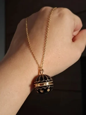 Svart och guldigt klotformat medaljonghalsband - Unikt halsband med klotformad medaljong i svart och guld. Medaljongen har detaljerade pärlkanter och kan öppnas för att förvara små saker. Kedjan är tunn och guldfärgad, vilket ger ett elegant intryck. Perfekt accessoar för dig som gillar vintagevibbar och detaljer.