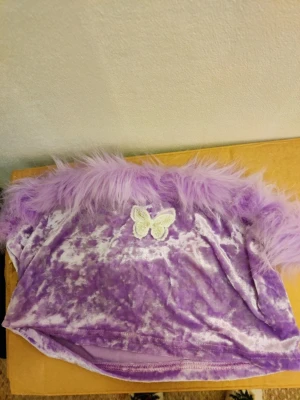 Lila tubtopp med fjädrar och fjäril - Superfin lila tubtopp i mjuk sammet med fluffiga fjädrar upptill och en söt broderad fjäril framtill. Perfekt statement-plagg för dig som vill sticka ut! Toppen är axelbandslös och har en croppad passform.