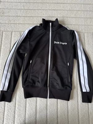 Svart track jacket från Palm Angels - Svart track jacket från Palm Angels med vita ränder längs ärmarna och vit dragkedja. Jackan har hög krage, två fickor med dragkedja och Palm Angels-logga tryckt på bröstet. Tillverkad i ett glansigt, sportigt material som passar perfekt till streetwear. Kvitto finns!! 