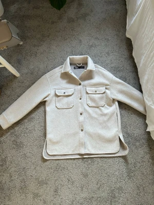 Ljusgrå overshirt från Zara - Stilren ljusgrå overshirt från Zara med två stora bröstfickor och knappar framtill. Overshirten har klassisk krage, långa ärmar och rundade slitsar i sidorna. Tillverkad i ett mjukt, filtliknande material som ger en clean och modern look. Använt bara ett par gånger.