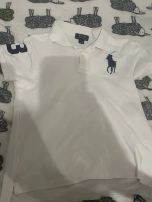 Vit pikétopp från Polo Ralph Lauren - Vit pikétopp från Polo Ralph Lauren med klassisk krage, knappar framtill och stor marinblå logga på bröstet. Siffran 3 i blått på ärmen ger en sportig känsla. Tillverkad i mjuk bomull och har korta ärmar.