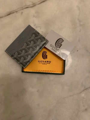 Grå kortfodral från Goy4ard - Snyggt grått kortfodral från Goy4ard med det klassiska mönstret i vitt och svart. Fodralet har flera fack för kort och är tillverkat i slitstarkt canvasmaterial med läderdetaljer. Perfekt för dig som vill ha en stilren och exklusiv accessoar.