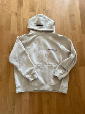 Beige Essentials hoodie  - Säljer en stilren beige hoodie från Essentials med svart tryck på bröstet och ärmen. Hoodien har en stor huva, magficka och ribbade muddar. Perfekt för en avslappnad och trendig look. Materialet känns mjukt och skönt mot huden.