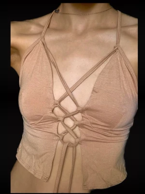 Beige croppad linnetopp med snörning - Trendig beige croppad linnetopp med smala axelband och snygg snörning framtill. Toppen har en omlottkänsla och är tillverkad i ett mjukt, stretchigt material som sitter tight mot kroppen. Perfekt för dig som gillar en modern och edgy stil.