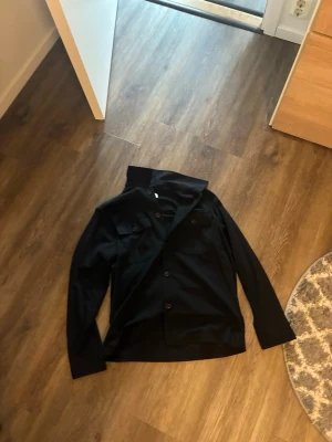 Svart overshirt från Selected Homme - Stilren svart overshirt från Selected Homme med klassisk krage och knäppning framtill. Två bröstfickor med lock ger en snygg och enkel look. Perfekt att bära över en t-shirt för en avslappnad men trendig stil.