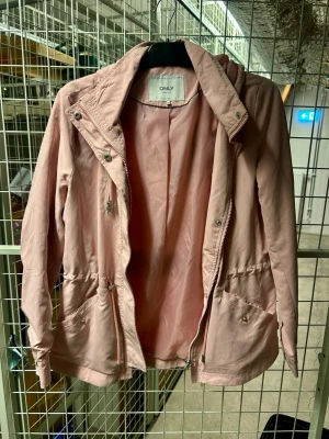 Ljusrosa vindjacka från ONLY - Snygg ljusrosa vindjacka från ONLY med dragkedja och tryckknappar framtill. Jackan har justerbar midja med snörning, två stora fickor med lock och knapp samt hög krage. Perfekt för blåsiga dagar och enkel att matcha till olika outfits.