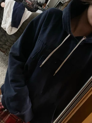 Mörkblå hoodie med vita snören GANT - Mysig mörkblå hoodie med huva och vita dragsnören. Klassisk känguruficka framtill och avslappnad passform. Perfekt för chill dagar och enkel att matcha med jeans eller mjukisbyxor. Materialet känns mjukt och bekvämt mot huden.