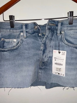 Blå jeanskjol från Zara - Trendig blå jeanskjol från Zara i klassisk femficksmodell. Kjolen har rå, fransig kant nertill och bälteshällor i midja. Stängs med knapp och dragkedja framtill. Oanvönd med lapp kvar. 