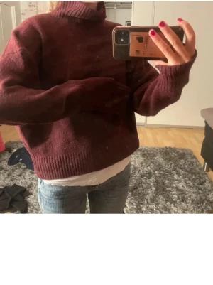 Vinröd stickad tröja med polokrage - Mysig vinröd stickad tröja med hög polokrage och långa ärmar. Tröjan har en avslappnad passform och ribbstickade muddar vid ärmslut och nederkant. Perfekt för kalla dagar och enkel att matcha med jeans.