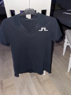 Marinblå pikétröja från J.Lindeberg - Klassisk marinblå pikétröja från J.Lindeberg med vit logga på bröstet. Tröjan har krage, korta ärmar och knappar vid halsen. Perfekt för en stilren och sportig look.