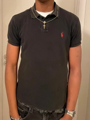 Svart pikétröja från Ralph Lauren - Klassisk svart pikétröja från Ralph Lauren med röd broderad logga på bröstet. Tröjan har korta ärmar, krage och knappar vid halsen. Tillverkad i mjuk bomull för en bekväm känsla och passar perfekt till jeans eller shorts. Det står att den är storlek M, men jag skulle säga att den mer är som  S.