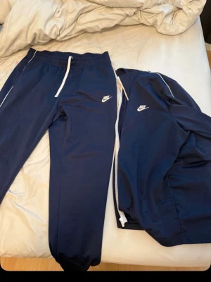Marinblå Nike träningsset - Säljer ett marinblått träningsset från Nike med vita detaljer. Setet består av en zip-jacka och matchande byxor, båda med Nike-logga på bröst och lår. Materialet är mjukt och stretchigt, perfekt för träning eller chill. Jackan har vita dragkedjor och piping längs ärmarna. Passar M