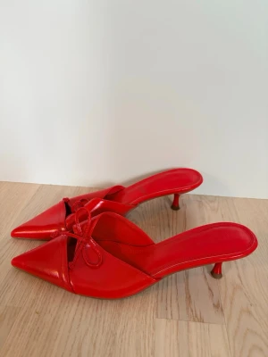 Röda spetsiga pumps med rosett - Snygga röda pumps med spetsig tå och låg klack. Skorna har en elegant rosettdetalj framtill och öppen häl. Tillverkade i glansigt material som ger en trendig look. Perfekta för dig som vill sticka ut med färg och stil.