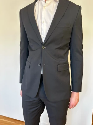 Svart kostym (inkluderar slips och sidenduk) - Stilren svart kostym med klassisk kavaj. Kavajen har två knappar, smala slag och fickor med lock. 