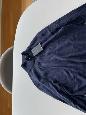 Mörkblå stickad tröja Tommy Hilfiger - Mörkblå stickad tröja från Tommy Hilfiger med halv dragkedja och liten broderad logga på bröstet. Tröjan har ribbstickad krage, ärmslut och nederkant. Perfekt för dig som gillar klassisk och stilren design.