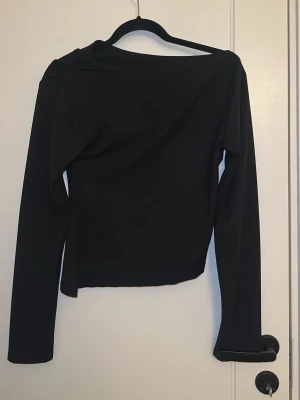 Svart asymmetrisk långärmad topp - Snygg svart långärmad topp med asymmetrisk axel och draperad effekt. Toppen har en unik skärning och sitter tight mot kroppen. Perfekt för dig som gillar minimalistisk och stilren design.