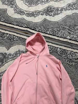 Ljusrosa hoodie från Polo Ralph Lauren - Säljer en ljusrosa hoodie från Polo Ralph Lauren med dragkedja framtill och klassisk blå logga broderad på bröstet. Hoodien har huva med dragsko och två fickor framtill. Tillverkad i mjukt bomullsmaterial för en skön och avslappnad look.