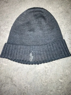 Mörkgrå stickad mössa Polo Ralph Lauren - Snygg mörkgrå stickad mössa från Polo Ralph Lauren med klassisk ribbad kant och broderad logga framtill. Perfekt för kalla dagar och enkel att matcha med olika outfits. Materialet är mjukt och värmande.