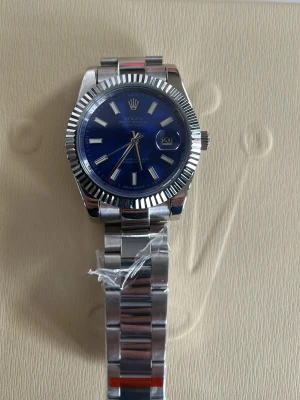 RLX klocka med blå urtavla - OBS automatiken funkar inte som den. Den går mestadels som den ska men ibland kan den haka upp sig ska därav pris. Endast klockan. Stilren klocka i rostfritt stål med blå urtavla och datumvisning. Klockan har en räfflad bezel och klassisk länkarmband i silverfärg. Perfekt accessoar för dig som gillar exklusiv och tidlös design.