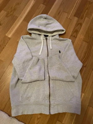 Raulph Lauren Hoodie - -Gråa hoodie med speciellt materiell vid huvan. Ett litet hål i huvan som syns i bilden, dragkedjan funkar galant! Längd:61cm Bredd: 50cm