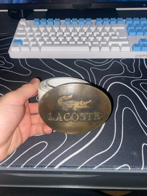 Vitt Lacoste bälte - Säljer ett vitt bälte från Lacoste med ett stort, ovalt metallspänne där Lacoste-loggan och krokodilen är ingraverade. Bältet har en clean look och passar perfekt till jeans eller byxor för att lyfta din outfit.