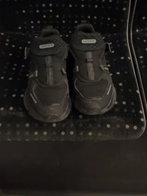 Svarta sneakers från Lacoste - Snygga svarta sneakers från Lacoste med meshdetaljer och reflekterande inslag. Skorna har en sportig design med robust sula och lågt skaft. Perfekta för dig som gillar streetwear och vill ha något stilrent men ändå funktionellt.