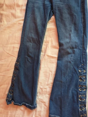 Blå bootcut jeans med snörning - Säljer ett par blå bootcut jeans med snygg snörningsdetalj längs bensluten. Jeansen har utsvängda ben och råa kanter längst ner för en trendig look. Perfekta för dig som gillar en unik och retro stil.